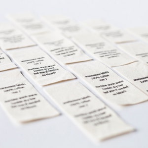 Op de afbeelding: Een close-up van verschillende witte stoffen labels met zwarte tekst. De labels lezen "handmade labels 100% cotton size 7" en "machine wash warm tumble dry low hot iron if needed no bleach".