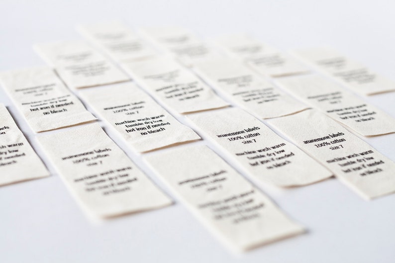 Op de afbeelding: Een close-up van verschillende witte stoffen labels met zwarte tekst. De labels lezen "handmade labels 100% cotton size 7" en "machine wash warm tumble dry low hot iron if needed no bleach".