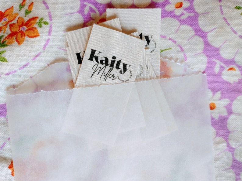 May include: A stack of white fabric tags with black text. The top tag reads "Kaity Miller Handmade Goods".