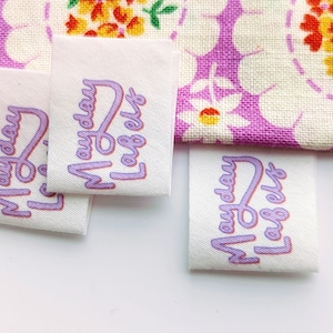 Puede incluir: Cuatro etiquetas de tela blanca con texto morado que dice "Monday Label" en una fuente estilizada.