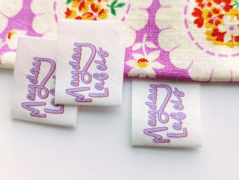Puede incluir: Cuatro etiquetas de tela blanca con texto morado que dice "Monday Label" en una fuente estilizada.