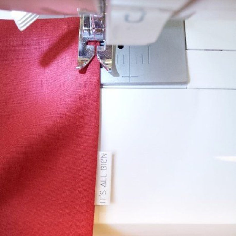 Puede incluir: Primer plano de una tela roja que se cose en una m&aacute;quina de coser blanca. La tela tiene una etiqueta blanca pegada que dice "IT'S ALL BIGN".