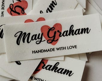 May include: A stack of white fabric labels with a red heart and the text "May Graham Handmade with Love" printed in black.