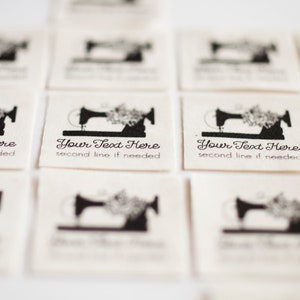 Peut inclure: Gros plan sur plusieurs &eacute;tiquettes en tissu blanc avec un motif de machine &agrave; coudre noire et le texte "Your Text Here" et "second line if needed".