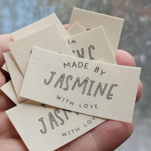 May include: A collection of rectangular, off-white fabric labels with black text. The labels read "MADE BY JASMINE WITH LOVE" in a handwritten style font. The labels are stacked and held in a hand.