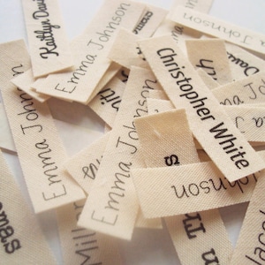 May include: A pile of off-white fabric labels with black ink printed names, including "Ethan Thomas", "Emma Johnson", "Christopher White", and "Jacob Miller".