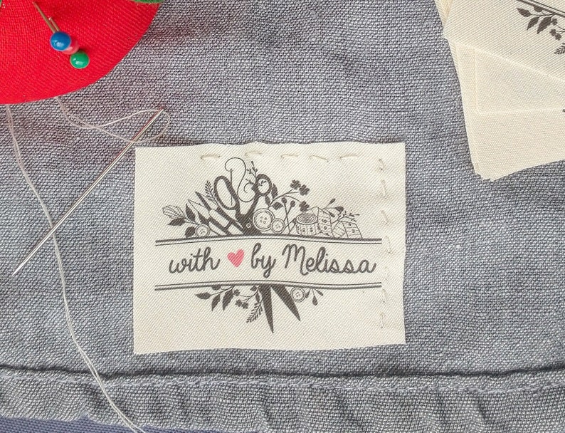 May include: A close-up of a fabric label with the text "with by Melissa" and a heart symbol. The label features a black and white illustration of sewing tools and floral elements. A red pin cushion with blue and green pins is in the background.