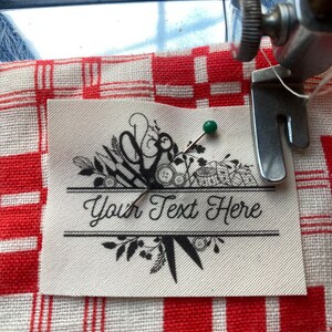 May include: A cream-colored label with the text "Your Text Here" is pinned to a red and white checkered fabric. The label features a decorative design with scissors, leaves, and other floral elements. A green pin secures the label.