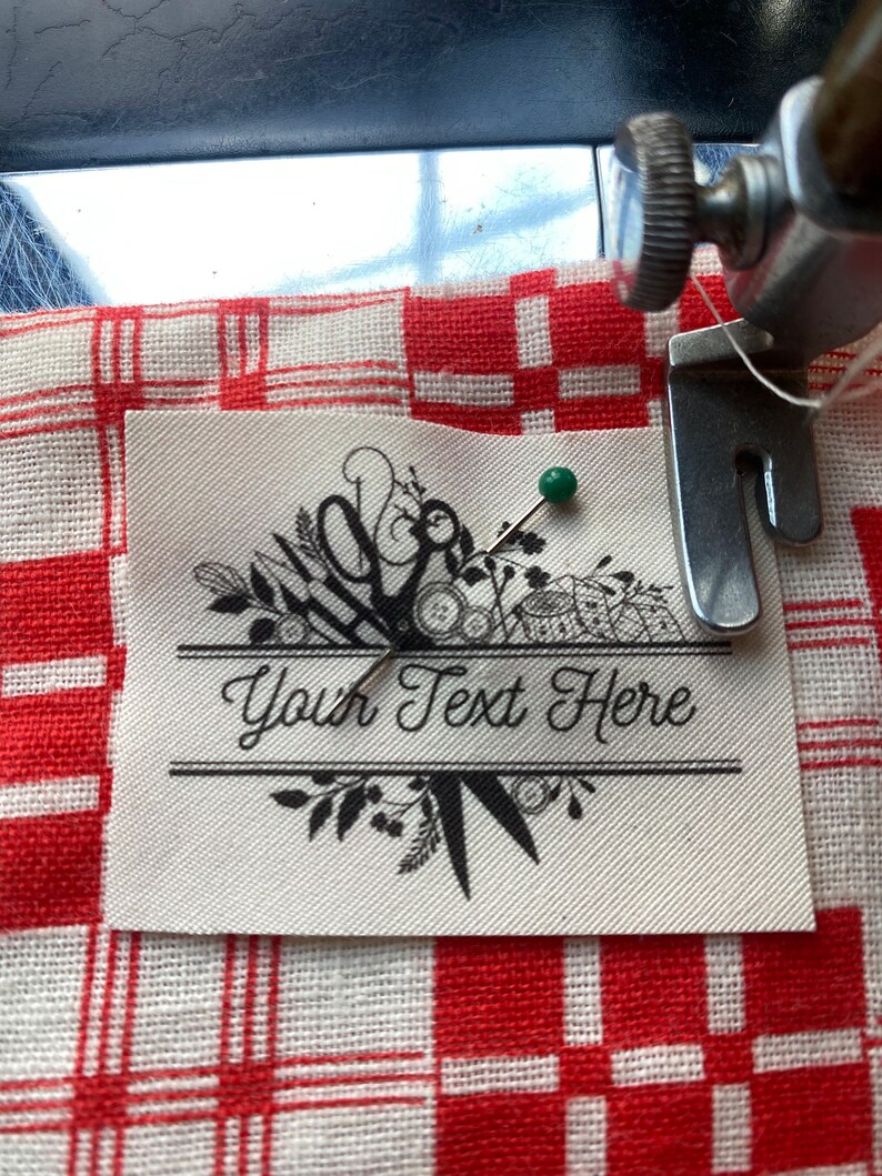 May include: A cream-colored label with the text "Your Text Here" is pinned to a red and white checkered fabric. The label features a decorative design with scissors, leaves, and other floral elements. A green pin secures the label.