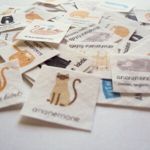 Puede incluir: Una colecci&oacute;n de peque&ntilde;as etiquetas cuadradas de tela con ilustraciones de gatos en naranja, marr&oacute;n, gris y negro. Cada etiqueta tiene el texto "ananemone labels" y algunas incluyen texto adicional como "custom, organic, handmade".