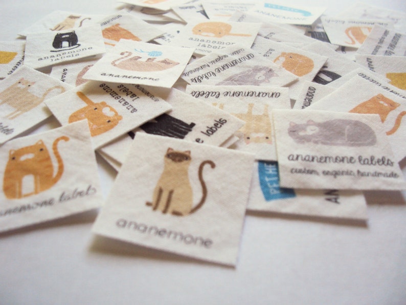 Puede incluir: Una colecci&oacute;n de peque&ntilde;as etiquetas cuadradas de tela con ilustraciones de gatos en naranja, marr&oacute;n, gris y negro. Cada etiqueta tiene el texto "ananemone labels" y algunas incluyen texto adicional como "custom, organic, handmade".