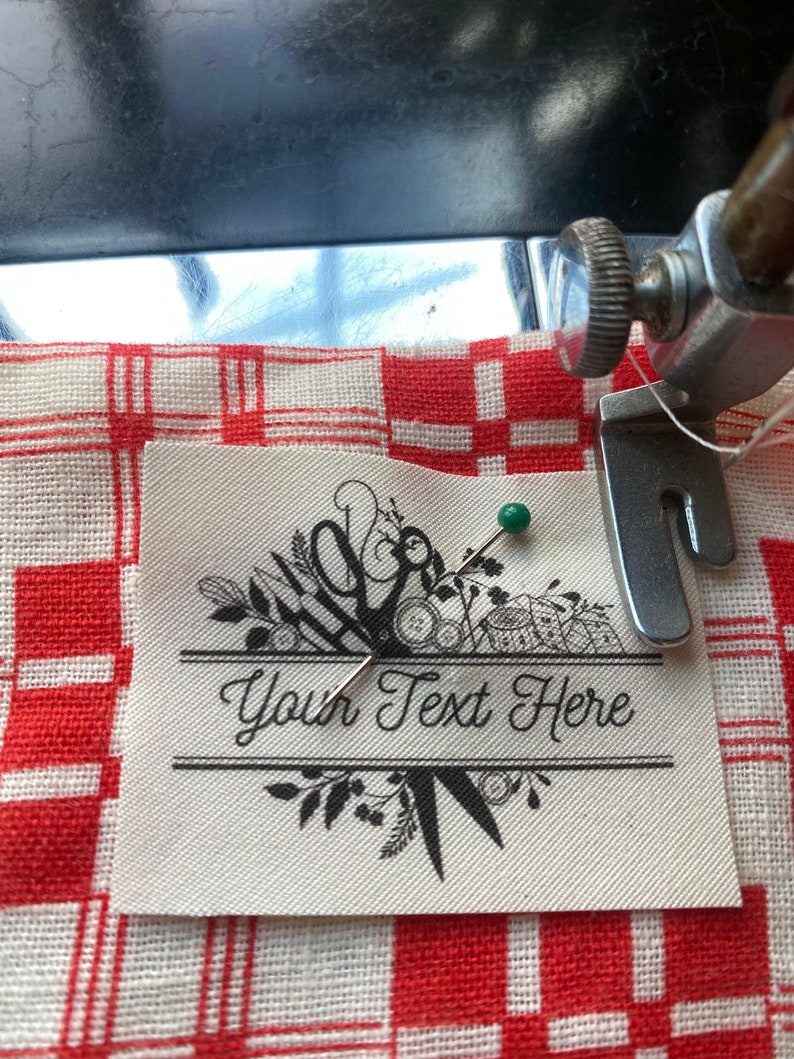 May include: A cream-colored fabric label with the text "Your Text Here" in a decorative font, framed by floral and scissor illustrations. A green pin secures the label to a red and white checkered fabric.