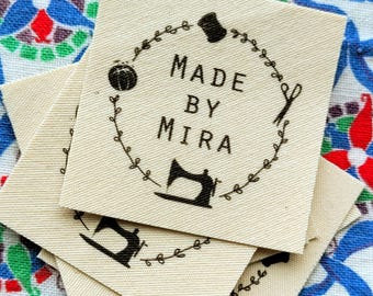 Op de afbeelding: Een stapel beige stoffen labels met een zwart cirkelontwerp. De cirkel bevat een naaimachine, een schaar, een spoel garen en de tekst "MADE BY MIRA".
