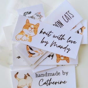 May include: A stack of white fabric labels with cat illustrations and text. The top label reads "Knit with love by Mandy" and the bottom label reads "Handmade by Catherine".