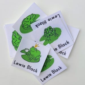 May include: A set of five white fabric labels with green frog illustrations and the text "Lewis Black" printed on them. Some of the frogs are wearing crowns.
