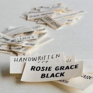 Può includere: Un primo piano di piccole etichette di tessuto bianco sporco con testo scritto a mano in nero. Le etichette sono sparse, alcune impilate e altre sparse. Un'etichetta mostra in modo prominente "ROSIE GRACE BLACK".