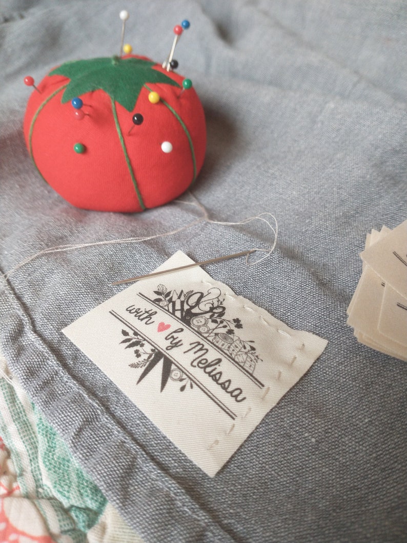 May include: A red tomato-shaped pin cushion with green leaves and various colored pins. A needle is threaded through a small fabric label that reads "with ❤️ by Melissa" with floral and sewing-themed illustrations. Additional labels are stacked nearby.
