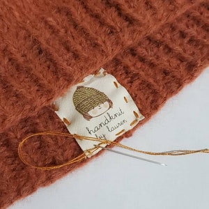 Puede incluir: Primer plano de un gorro de punto marr&oacute;n con una etiqueta blanca cosida. La etiqueta presenta una ilustraci&oacute;n de dibujos animados de una persona con un sombrero y el texto "handknit by Lauren".