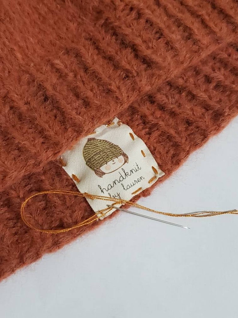 Puede incluir: Primer plano de un gorro de punto marr&oacute;n con una etiqueta blanca cosida. La etiqueta presenta una ilustraci&oacute;n de dibujos animados de una persona con un sombrero y el texto "handknit by Lauren".