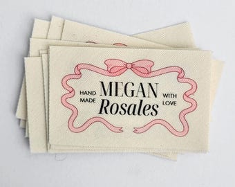 May include: A stack of white fabric labels with pink ribbon and bow design. The text on the label reads "Hand Made Megan Rosales With Love".