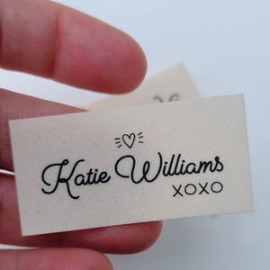 Puede incluir: Una etiqueta rectangular de tela blanquecina con el nombre "Katie Williams" en escritura cursiva negra, un símbolo de corazón y las letras "XOXO". La etiqueta se sostiene entre los dedos.