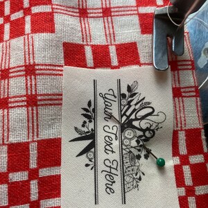 May include: A sewing machine needle is positioned over a fabric label with the text "Your Text Here". The label features a black floral and scissor design. The fabric is pinned to a red and white checkered cloth.