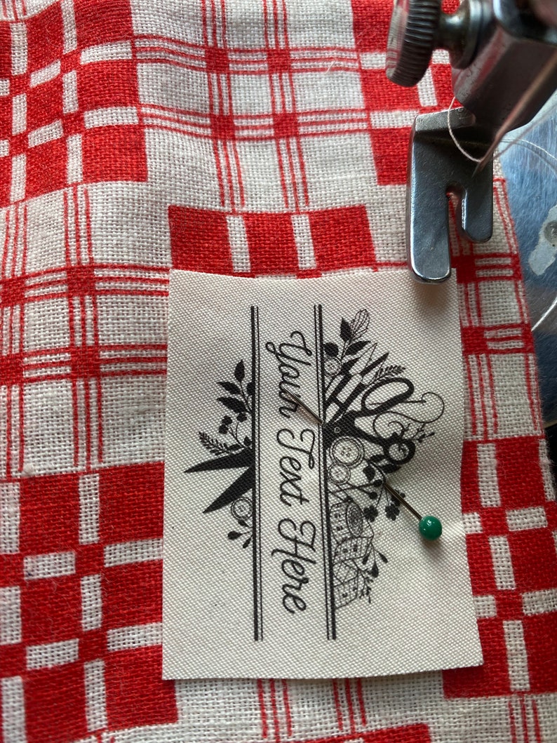 May include: A sewing machine needle is positioned over a fabric label with the text "Your Text Here". The label features a black floral and scissor design. The fabric is pinned to a red and white checkered cloth.