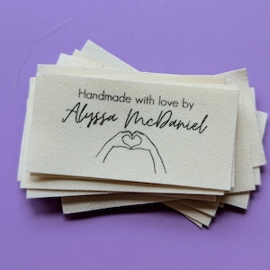 May include: A stack of white fabric labels with black text that reads "Handmade with love by Alyssa McDaniel" and a hand drawn heart shape.