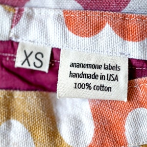 May include: A white fabric label with black text that reads "anemone labels handmade in USA 100% cotton". A smaller white fabric label with black text that reads "XS" is visible above the larger label.