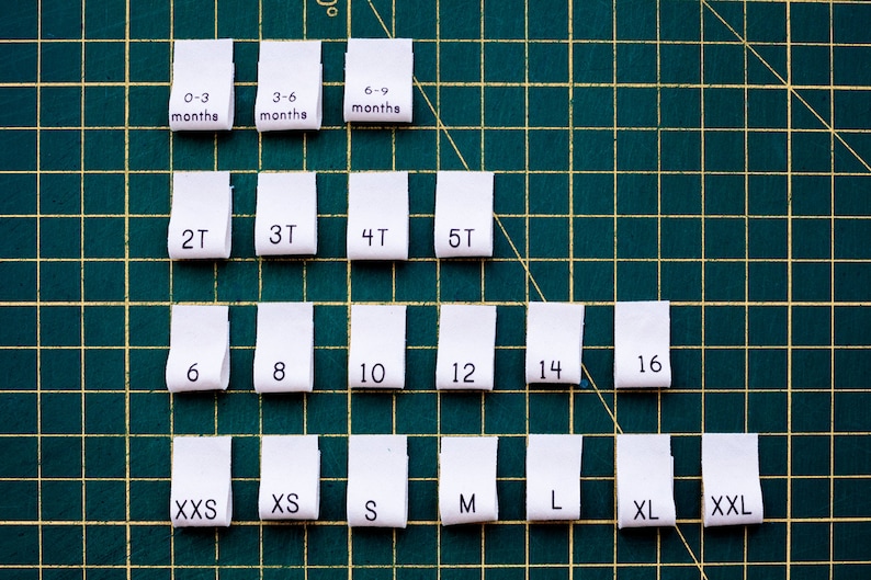 May include: White fabric labels with different sizes for clothing, including 0-3 months, 3-6 months, 6-9 months, 2T, 3T, 4T, 5T, 6, 8, 10, 12, 14, 16, XXS, XS, S, M, L, XL, and XXL. The labels are arranged in a pyramid shape on a green cutting mat.