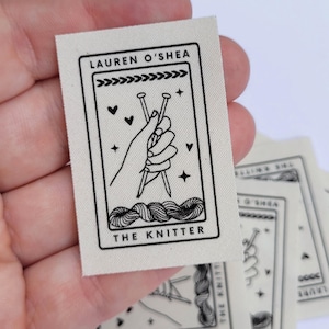 Pode incluir: Um r&oacute;tulo de tecido branco com texto preto e um gr&aacute;fico. O texto diz "Lauren O'Shea" e "The Knitter". O gr&aacute;fico &eacute; uma m&atilde;o segurando agulhas de tric&ocirc; e fio.
