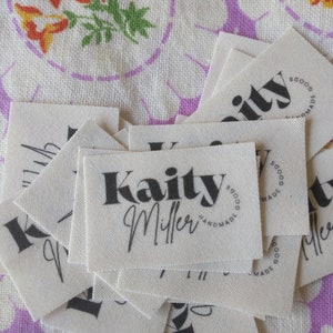 May include: A stack of white fabric labels with black text. The top label reads "Kaity Miller" with a circle around the text that says "Handmade Goods".