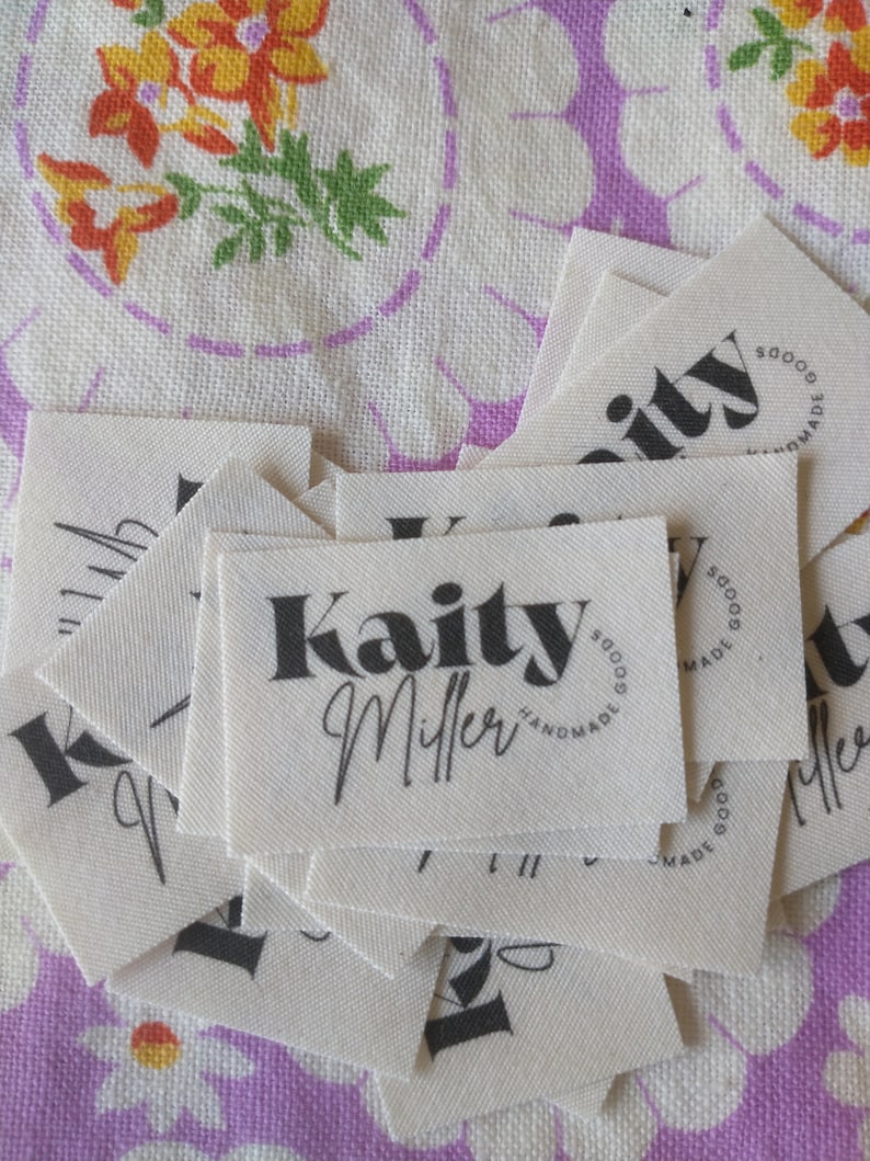 May include: A stack of white fabric labels with black text. The top label reads "Kaity Miller" with a circle around the text that says "Handmade Goods".