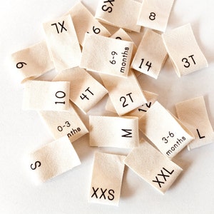 May include: A collection of beige fabric clothing size tags with black text. The tags include sizes such as XS, S, M, L, XL, XXL, XXXS, 0-3 months, 3-6 months, 6-9 months, 2T, 3T, 4T, 7X, 9, 10, 12, 14, and 8.