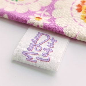 Puede incluir: Etiqueta de tela blanca con el texto "Mayday Labels" impreso en morado.