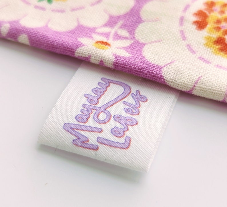 Puede incluir: Etiqueta de tela blanca con el texto "Mayday Labels" impreso en morado.