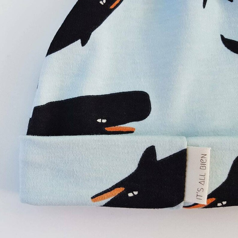 Puede incluir: Un gorro azul claro con un patr&oacute;n de ballena negra y el texto "IT'S ALL BIG'N" en una etiqueta blanca.