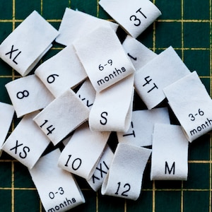 May include: A collection of white fabric clothing size tags with black text. The tags include sizes XL, 8, 6, 3T, 6-9 months, 14, LXS, S, 10, 4T, 3-6 months, 12, M, 0-3 months, AXI.