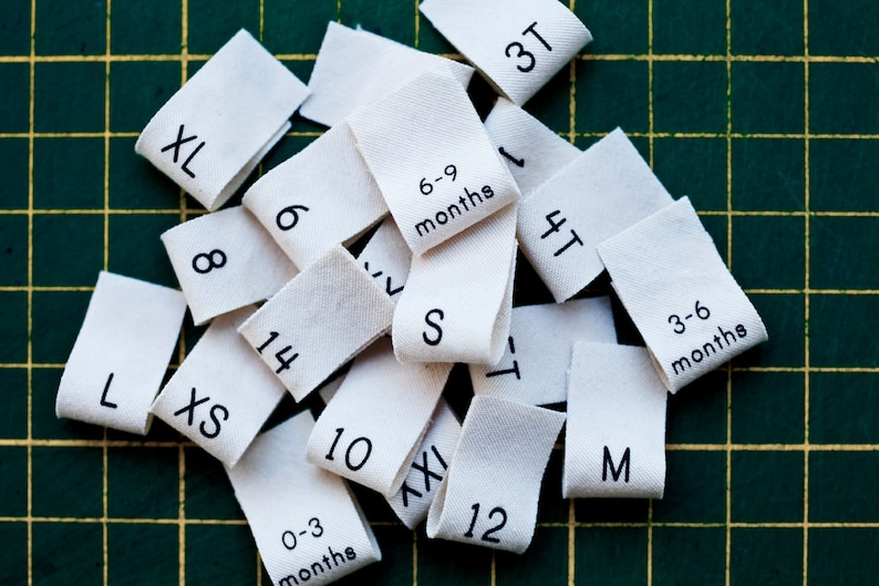 May include: A collection of white fabric clothing size tags with black text. The tags include sizes XL, 8, 6, 3T, 6-9 months, 14, LXS, S, 10, 4T, 3-6 months, 12, M, 0-3 months, AXI.