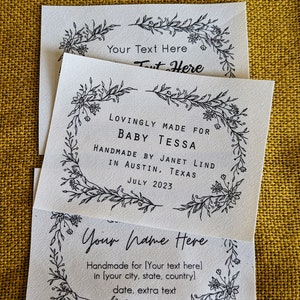 May include: Three white labels with black floral borders and text. The top label says "Your Text Here Text Here". The middle label says "Lovingly Made For Baby Tessa Handmade by Janet Lind in Austin, Texas July 2023". The bottom label says "Your Name Here Handmade for [Your text here] in [your city, state, country] date, extra text".