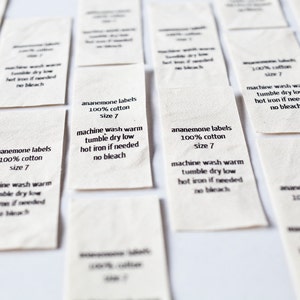 Op de afbeelding: Een verzameling witte stoffen labels met zwarte tekst. De labels lezen "anemone labels 100% cotton size 7" en bevatten wasinstructies: machinewas warm, drogen in de droger op lage temperatuur, heet strijken indien nodig, geen bleekmiddel.