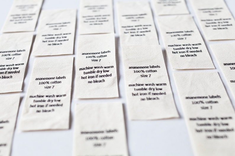 Op de afbeelding: Een verzameling witte stoffen labels met zwarte tekst. De labels lezen "anemone labels 100% cotton size 7" en bevatten wasinstructies: machinewas warm, drogen in de droger op lage temperatuur, heet strijken indien nodig, geen bleekmiddel.