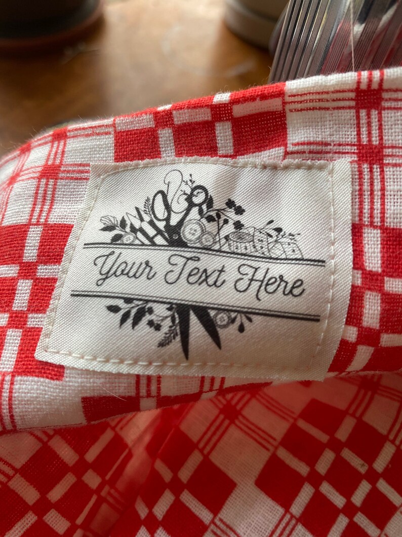 May include: Close-up of a red and white checkered fabric with a white label. The label features the text "Your Text Here" in elegant script, framed by floral and scissor illustrations. The fabric has a classic picnic blanket pattern.