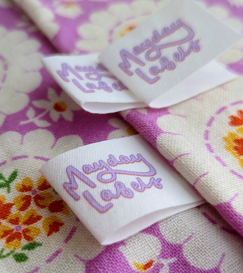 Puede incluir: Tres etiquetas de tela blanca con el texto "Mayday Labels" en letras moradas. Las etiquetas est&aacute;n sobre una tela floral rosa y blanca.