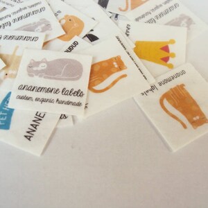 Puede incluir: Un conjunto de etiquetas de tela blanca con ilustraciones de gatos en naranja, gris y negro. Las etiquetas incluyen el texto "ananemone labels" y "custom, organic, handmade."