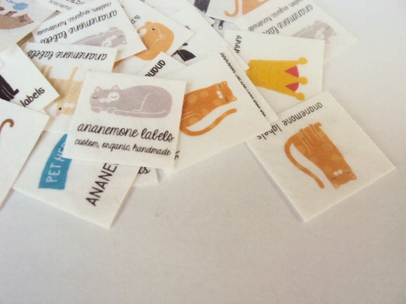 Puede incluir: Un conjunto de etiquetas de tela blanca con ilustraciones de gatos en naranja, gris y negro. Las etiquetas incluyen el texto "ananemone labels" y "custom, organic, handmade."