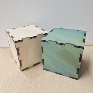 May include: Two wooden cube boxes. One is natural wood color, the other is painted with shades of blue and green. Both have dark brown corner joints. The boxes are on a light wood surface.