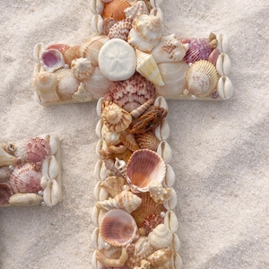 May include: A decorative cross made of various seashells, including conch shells, scallops, and sand dollars. The shells are in shades of white, beige, pink, and brown, arranged on a sandy surface.