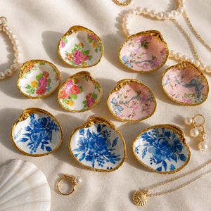 May include: A collection of eight decorative clam shells with gold-painted edges, featuring interior floral patterns in pink, yellow, and blue. The shells are arranged alongside pearl necklaces, gold rings, and a small gold shell pendant.