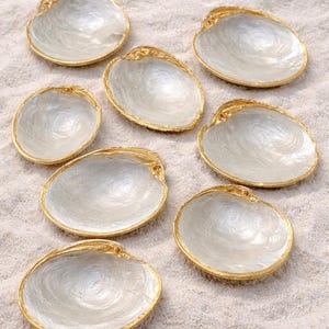 Puede incluir: Conjunto de ocho conchas decorativas con interior nacarado y borde dorado. Los platos están dispuestos sobre arena, con algunas conchas en el fondo. Ideales para decoración o servir pequeñas porciones.